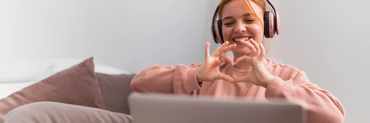 Frau mit Kopfhörern zeigt in einem Online-Meeting ein Herzzeichen mit den Händen – Symbol für digitale Nähe und positive Online-Kommunikation.